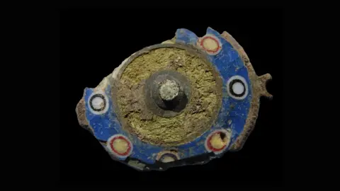 A Roman brooch on a black background. The brooch is oval shaped, but has parts missing on its sides. It is a dark yellow colour in the centre. Around the outside, it is blue with blue and white circles and red and yellow circles.