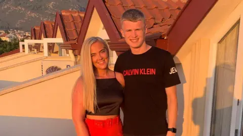 SWNS A man and a woman posing side by side for a photograph outside some lodges in a sunny place. The woman is blonde, wearing a black top and red trousers and has a silver bracelet on her right hand. The man is wearing a black top and shorts. 
