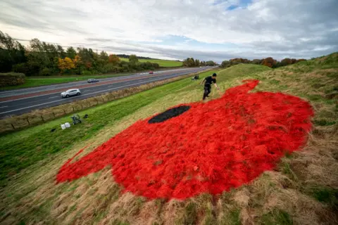 Jane Barlow/PA Media A motorway cuts through a countryside landscape of trees and fields.  On a raise area of grass to the right and man in black uses red paint to create a picture of a remembrance poppy on the ground 