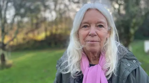 Jacqi Hodgson smiles at the camera, she is wearing a pale green coat and a pink scarf. She has long white hair. 