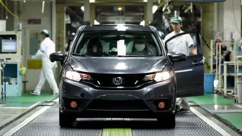 A grey Honda car seen in a factory in Japan. A factory workers in white overalls can be seen opening the door and another factory worker in the background 
