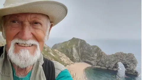 Derek Adams Derek Adams with short white hair and beard, wearing a light-coloured hat with a wide brim. He is looking at the camera with a coastline behind him, showing green and grey rocks and a sand path. There is a natural stone arch above the water to the right.
