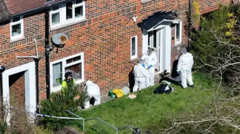 Imagen aérea de cuatro personas vestidas con trajes forenses blancos afuera de una casa adosada.