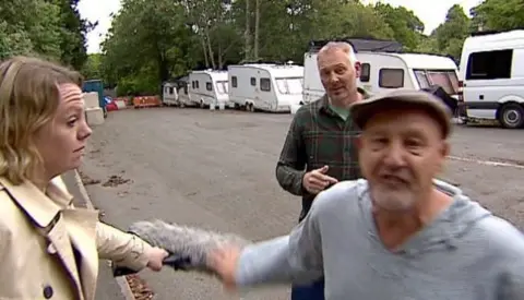 Man shouting at camera, with reporter and another man stood in the background
