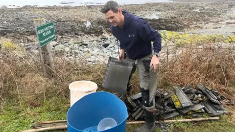 A Man in a blue hoodie and grey trousers, picking up broken rood tiles and putting them in a blue plastic tub