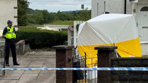 The crime scene at Rocky Road Merthyr Tydfil. There's a forensic tent outside a white house with a white garden wall. A police car is on the left hand side and there's police tape in front of the house.