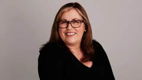 Mid-shot of Liza Tarbuck wearing a black top, leopard print frames and wearing streaks of blonde in her long flowing hair.