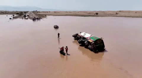     Reuters Mensen staan ​​naast een omgevallen voertuig in het overstromingswater in Jemen. 