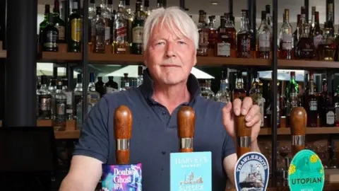 Mark Dorber a man looking directly at the camera and smiling. He is standing inside his pub, The Swan, behind the bar and is wearing a dark blue T-shirt.