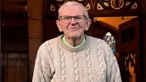 GERRY CALLISTER Gerry, un hombre de pelo blanco y grandes gafas de lectura, viste un jersey gris y aparece en la imagen dentro de una iglesia.