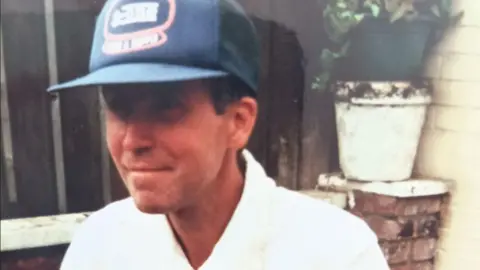 Norfolk Police Maris Ile wears a white shirt and a baseball-type blue hat. he is sat in a courtyard with a flower pot behind him.
