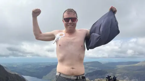 A topless man wearing sunglasses with his arms raised in the air and a t-shirt in one hand with a view from a hilltop behind him and a white medical patch on his chest.