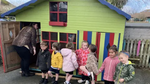 A bright lime coloured wooden playhouse with wed painted windows and red, lime green and blue doors stands in the playground with seven children standing in front all smiling, some have turned around. The teacher is inside the playhouse trying to coax a small child out 