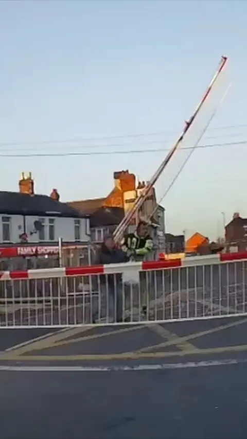 A man in hi-vis pulls an elderly man in dark clothes from behind level crossing barriers.