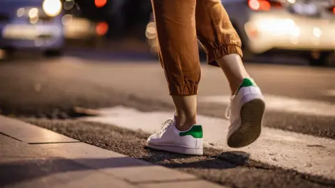 A women's legs in brown trousers and white trainers walking on a pavement at night