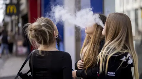 Three young girls blow huge plumes of aerosol mist from their vapes. They appear to be stood on a high street. 