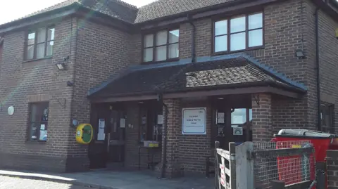 Google The exterior of the Higham surgery of the Highparks Medical Practice, a dark red brick modern building, with a wooden gate entrance and a metal wire fence.