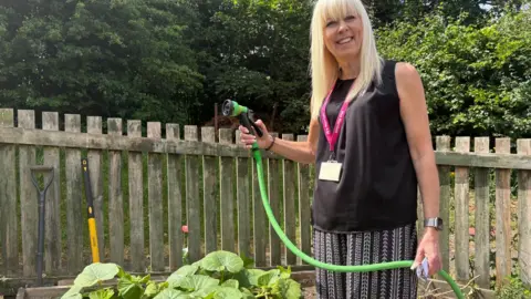 Simon Thake/BBC A woman with a blonde fringe stands in an allotment holding a green hose.