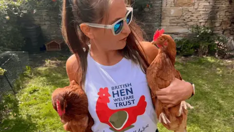 A woman with long, brown hair and white sunglasses with blue lenses holding a brown chicken under each arm. She is wearing a shirt which says "British Hen Welfare Trust".