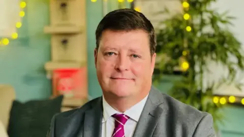 Head-and-shoulders photo of Sir Martyn Oliver, a middle-aged man in a grey suit with a pink tie