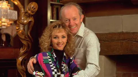Debbie McGee, wearing a colourful outfit, with her late husband Paul Daniels in 1992. They are sat close to each other and Paul has his arms around her.