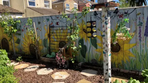 Shariqua Ahmed/BBC A yellow sunflower mural painted on a white fence. 