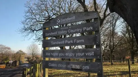 Local Democracy Reporting Service A mainly dark sign that includes the words Welcome to Sandwell Valley Country Park. Trees and grass are in the background.