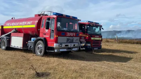 HFRS/Pocklington Fire Station A red water bowser, with a large tank on the back of an HGV, next to a fire engine overlooking fields with smoke in the distance.