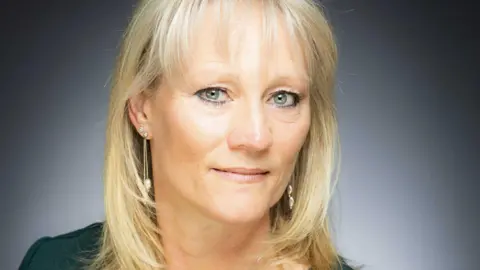 Sharon Hadley GP Sharon Hadley looks at the camera. She has blonde hair and grey eyes with light pink lipstick. She has dangly silver earrings in and is wearing a dark green top