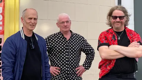 Adrian Goldberg Three men stand in front of a white brick wall. The man on the left has grey hair and is wearing a blue jacket. The man in the centre has grey hair and glasses, and is wearing a black and white polka dot shirt. The man on the right has dark blonde hair and a beard, and sunglasses, and is wearing a red shirt.