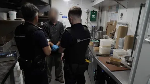 Inside a kitchen area showing the backs of two immigration officers in black uniforms, talking to a man whose face is blurred - he is wearing chef top in black and has blue food prep gloves on. Either side are stainless steel worktops with food being prepared.