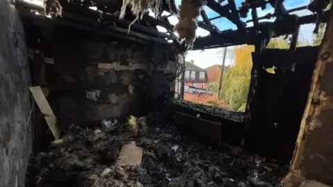 ECFRS A bedroom on the upper floor of a house. The window is missing and fire has torn through the roof so that the sky is visible through the remaining rafters. The entire room is destroyed and black and insulation from the loft is littering the floor and hanging from the charred roof timbers.