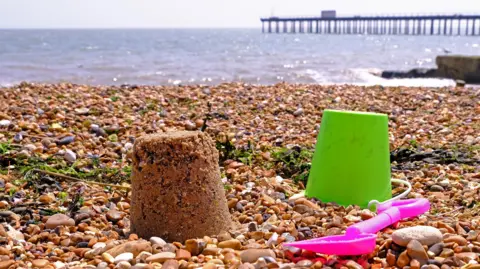 A sandcastle rests on a shingle beach. A green bucket and pink space rest next to it. A pier can be seen in the background leading into the sea.