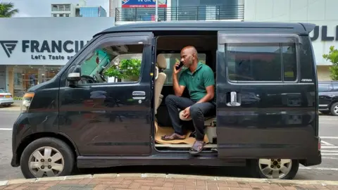 BBC Ajeewan Sadasivam is seen on the phone, perched in the doorway of his black minivan cab