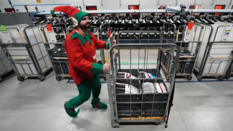 PA Media A man dressed as Christmas elf pushes a trolley of letters to Santa in a Royal Mail sorting office. He is wearing a green and red tunic, green and red pointy hat with large elf ears and pointy-toed green shoes. 