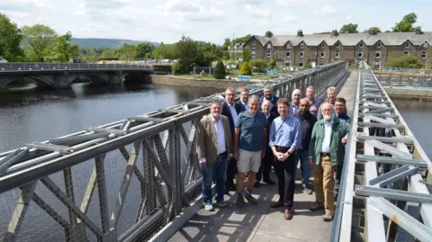 A group of people stand on a footbridge, which runs over a river, on a sunny day. In the background are some houses and another bridge.