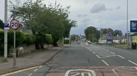 Google A high street with the entrance to a supermarket on one side and a trees on the other. A sign says the speed limit is 20mph