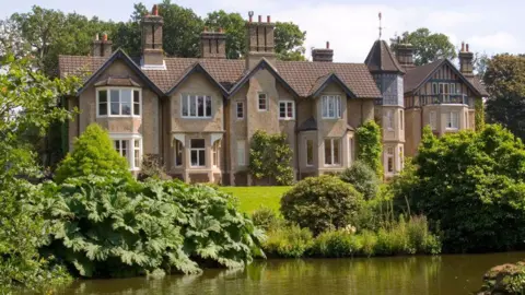 Alamy York Cottage on the Sandringham estate. It is a large, brown-brick two-floor property set near a lake. It has a brown roof and a turret on the right-hand side.