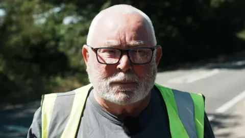 Tony Maher is wearing a grey T-shirt and a hi-vis vest. He has short grey hair, a beard and glasses. He looks serious in his expression, standing next to a road.