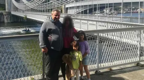 Bartlomiej Kuczynski, who has grey hair and is wearing a grey hoodie, stands with his arm round a woman wearing a red top. Her face is blurred. Next to them are Jasmin Kuczynska and Natasha Kuczynska. They are standing beside two bridges across a river.