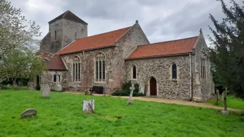 Geograph/David Bremner Exterior of All Saints Church in Ashwicken, King’s Lynn, a stone and flint building with ornate arched windows with trees either side and gravestones in front