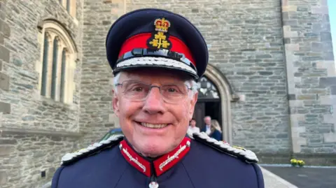 A man, wearing a cap and a navy uniform jacket, smiling at the camera. There is a cathedral building behind him.