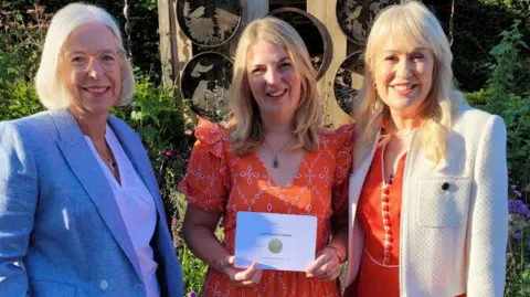 Three women next to each other smiling with the woman in the middle holds a card showing her silver gilt winning medal