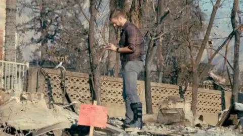 A man with a leg in a cast looks at the remnants of his house after a fire