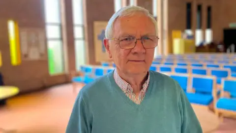 SHARIQUA AHMED/BBC Philip stands wearing a blue v-neck jumper and a checked shirt beneath. He is standing inside the main prayer hall of the church, with blue chairs in the background