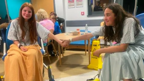 The girls in hospital gowns with blankets over legs and in hospital chairs on a ward reaching out to hold each other's hand