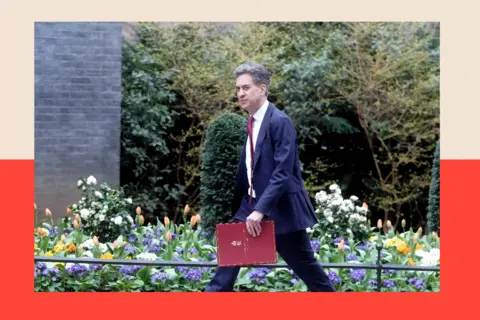 PA Media Ed Miliband walking in Downing Street, carrying a red briefcase. There are slpring flowers in bloom behind him, including orange tulips.