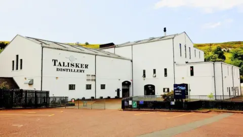 The distillery is a white-walled building with long rectangle windows. The words "Talisker Distillery" is on one wall.