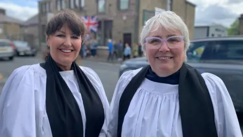 BBC/Phil Cunliffe Two women vicars dressed in white vestments stand smiling in a street.