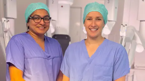 Two women wearing blue scrubs and green hair nets. They are smiling at the camera and standing in a medical room